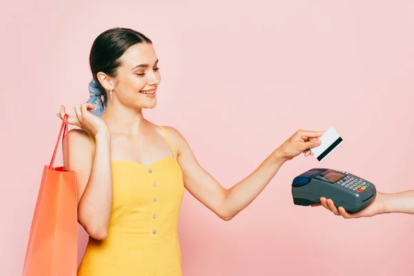 Brunette jeune femme avec sac à provisions payer avec carte de crédit isolé sur rose — Photo de stock