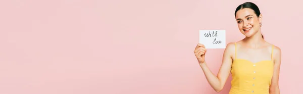 Morena joven sosteniendo con tarjeta de amor aislado en rosa, tiro panorámico - foto de stock