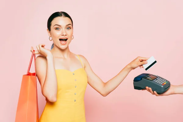 Chocado morena jovem com saco de compras pagando com cartão de crédito em rosa — Fotografia de Stock
