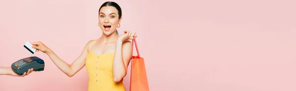 Choqué jeune femme brune avec sac à provisions payer avec carte de crédit sur rose, plan panoramique — Photo de stock