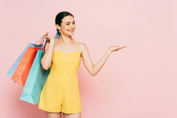 Brünette junge Frau mit Einkaufstaschen, die mit der Hand zur Seite auf rosa zeigen — Stockfoto