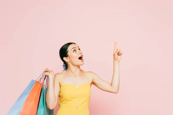 Chocado morena jovem com sacos de compras apontando para cima em rosa — Fotografia de Stock