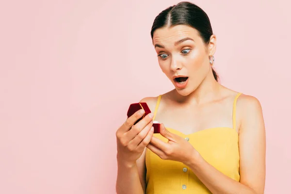 Chocado morena jovem segurando caixa com anel de noivado isolado em rosa — Fotografia de Stock