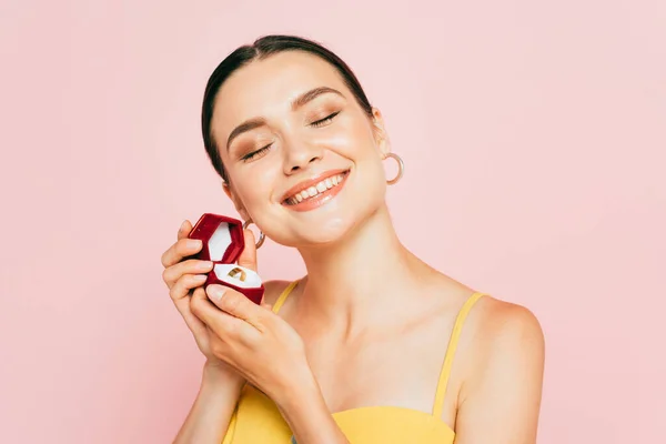 Задоволена брюнетка молода жінка з закритими очима тримає коробку з заручинним кільцем ізольовано на рожевому — стокове фото