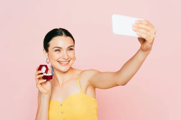 Foco seletivo de morena jovem segurando caixa com anel de noivado enquanto toma selfie isolado no rosa — Fotografia de Stock