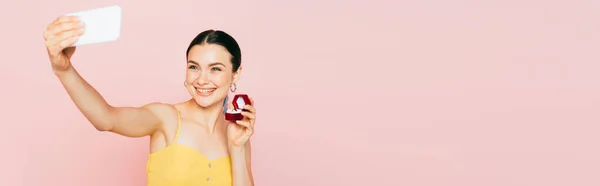 Foyer sélectif de la jeune femme brune tenant boîte avec bague de fiançailles tout en prenant selfie isolé sur rose, plan panoramique — Photo de stock