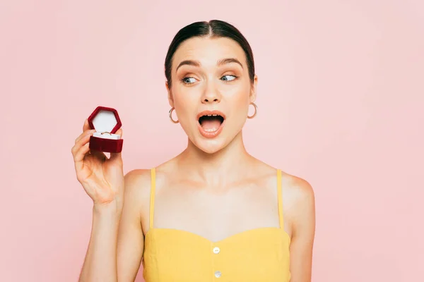 Shocked brunette young woman holding box with engagement ring isolated on pink — Stock Photo