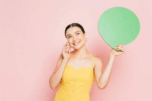 Brunette young woman with green blank speech bubble talking on smartphone on pink — Stock Photo