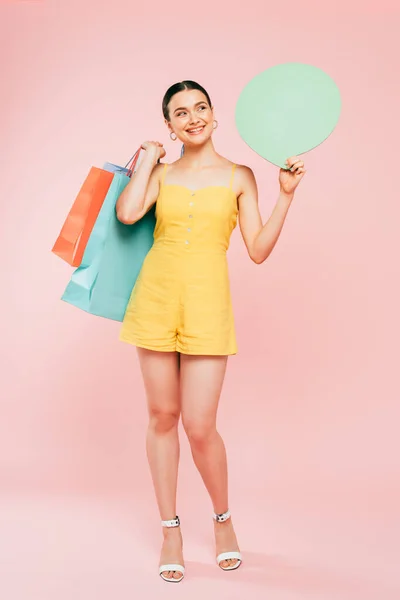 Повнометражний вид на брюнетку молоду жінку з сумками для покупок і мовою бульбашки на рожевому — стокове фото