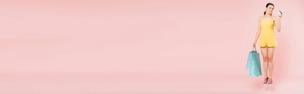 Vista completa de salto morena joven con bolsas de compras y tarjeta de crédito en rosa, plano panorámico - foto de stock