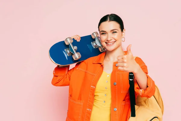 Brunette young woman in colorful outfit with skateboard showing thumb up isolated on pink — Stock Photo