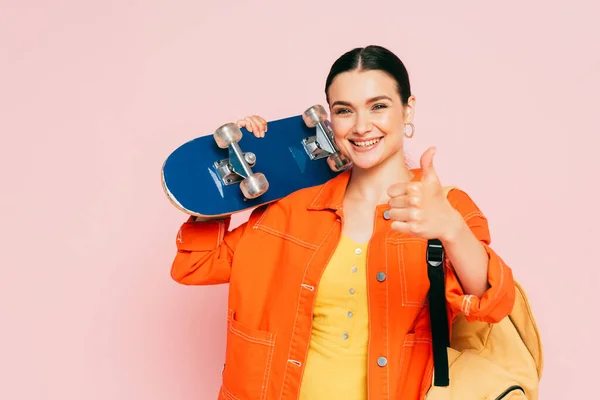 Brunetta giovane donna in abito colorato con skateboard mostrando pollice in su isolato su rosa — Foto stock