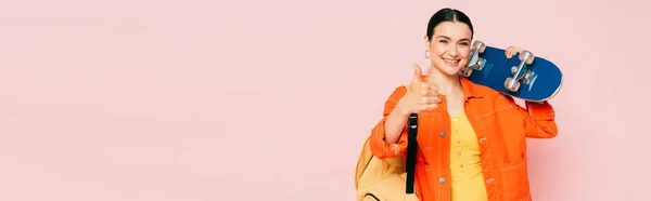 Brunette jeune femme en tenue colorée avec planche à roulettes montrant pouce vers le haut isolé sur rose, vue panoramique — Photo de stock
