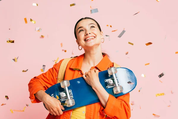 Brunette young woman in colorful outfit with skateboard under falling confetti isolated on pink — Stock Photo