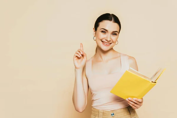 Morena joven con libro amarillo mostrando gesto de idea aislado en beige - foto de stock