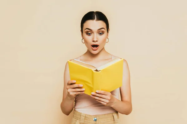 Chocado morena jovem leitura amarelo livro isolado no bege — Fotografia de Stock