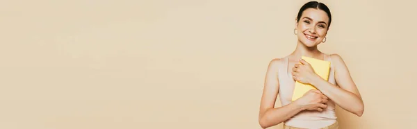 Brunette young woman hugging yellow book isolated on beige, panoramic shot — Stock Photo