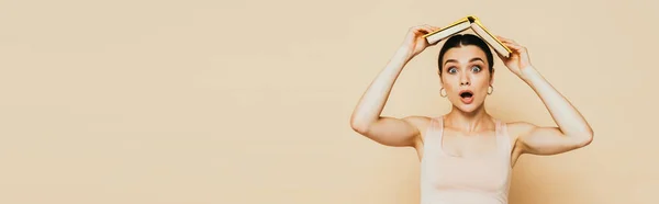 Shocked brunette young woman with book on head on beige, panoramic shot — Stock Photo