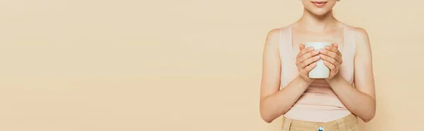 Обрізаний вид молодої жінки, що тримає білий кухоль ізольовано на бежевому, панорамному знімку — стокове фото