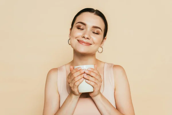 Pleased brunette young woman with closed eyes holding mug isolated on beige — Stock Photo