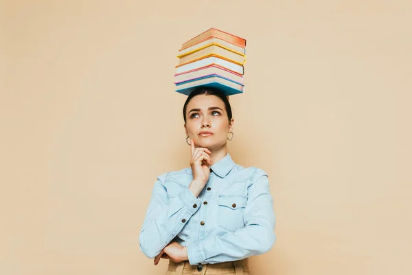 Pensive student in denim shirt with books on head isolated on beige — Stock Photo