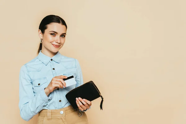 Brunette woman in denim shirt holding wallet and credit card isolated on beige — Stock Photo