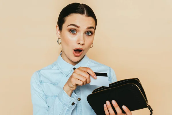 Shocked brunette woman in denim shirt holding wallet and credit card isolated on beige — Stock Photo