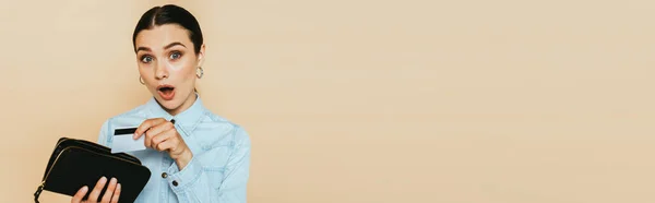 Shocked brunette woman in denim shirt holding wallet and credit card isolated on beige, panoramic shot — Stock Photo