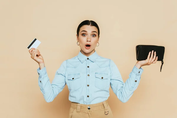Schockierte brünette Frau im Jeanshemd mit Geldbörse und Kreditkarte isoliert auf beige — Stockfoto