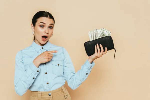 Shocked brunette woman in denim shirt pointing at wallet with dollars isolated on beige — Stock Photo