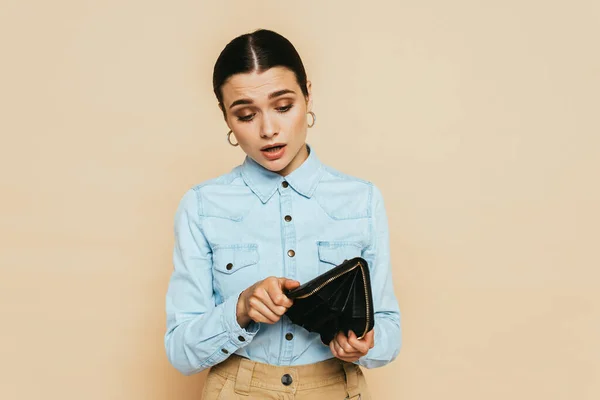 Sad brunette woman in denim shirt holding empty wallet isolated on beige — Stock Photo