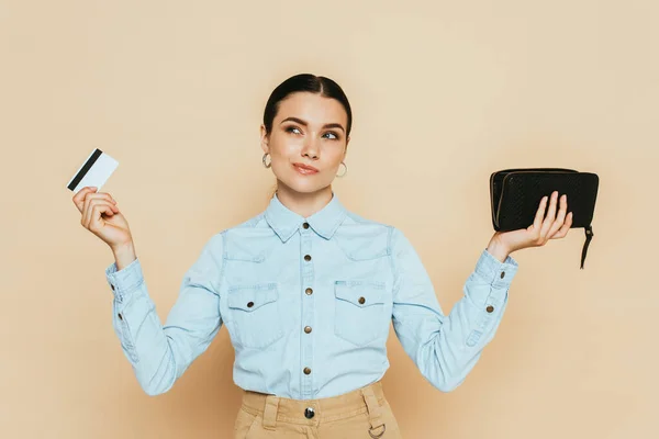 Pensive brunette woman in denim shirt holding wallet and credit card isolated on beige — Stock Photo