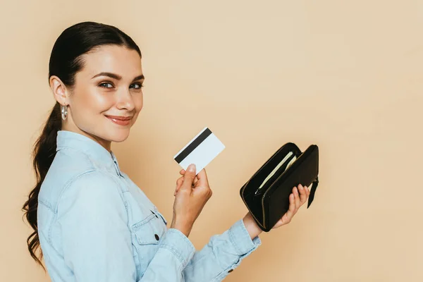Side view of brunette woman in denim shirt holding wallet and credit card isolated on beige — Stock Photo