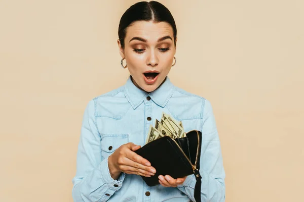Shocked brunette woman in denim shirt holding wallet with dollars isolated on beige — Stock Photo