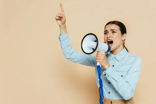 Brunette woman in denim shirt screaming in loudspeaker on beige — Stock Photo