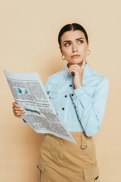 Pensive brunette woman in denim shirt with business newspaper on beige — Stock Photo