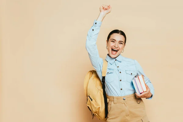 Aufgeregte brünette Studentin im Jeanshemd mit Büchern und Rucksack auf beige — Stockfoto