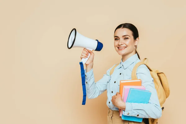 Side view of brunette student in denim shirt with books and backpack holding loudspeaker on beige — Stock Photo