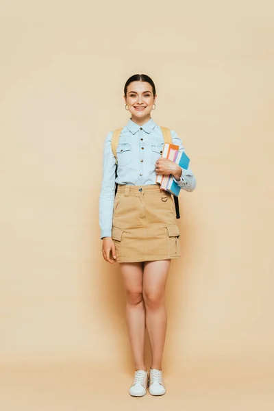 Vista completa de estudiante morena en camisa de mezclilla con libros y mochila en beige - foto de stock