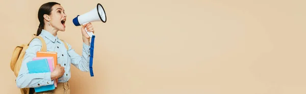 Side view of brunette student in denim shirt with books and backpack shouting in loudspeaker on beige, panoramic shot — Stock Photo