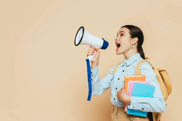 Side view of brunette student in denim shirt with books and backpack shouting in loudspeaker on beige — Stock Photo