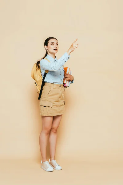 Full length view of brunette student in denim shirt with books and backpack pointing with finger on beige — Stock Photo