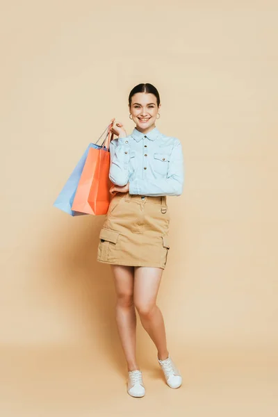 Vista a figura intera di donna bruna in camicia di jeans con borse della spesa su beige — Foto stock
