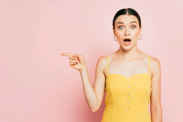 Schockierte brünette junge Frau zeigt zur Seite auf rosa — Stockfoto