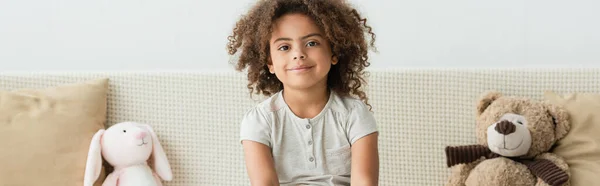 Panoramic crop of curly african american kid looking at camera near soft toys — Stock Photo