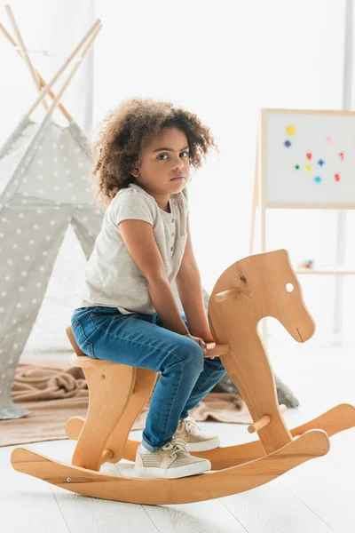 Curly african american kid sitting on wooden rocking horse at home — Stock Photo