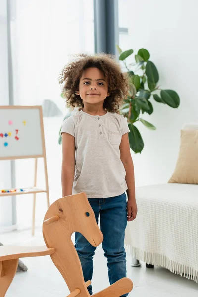 Curly african american standing near wooden rocking horse — Stock Photo