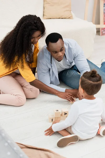 Избирательный фокус африканских американских родителей, смотрящих на ребенка, сидящего на полу рядом с деревянными кубиками — стоковое фото