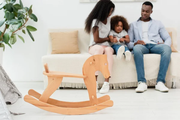 Focus selettivo del cavallo a dondolo in legno vicino alla famiglia afroamericana seduta sul divano — Foto stock