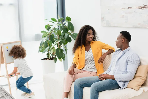 Enfoque selectivo de los padres afroamericanos sentados en el sofá, mientras que la hija de pie cerca del caballete magnético - foto de stock